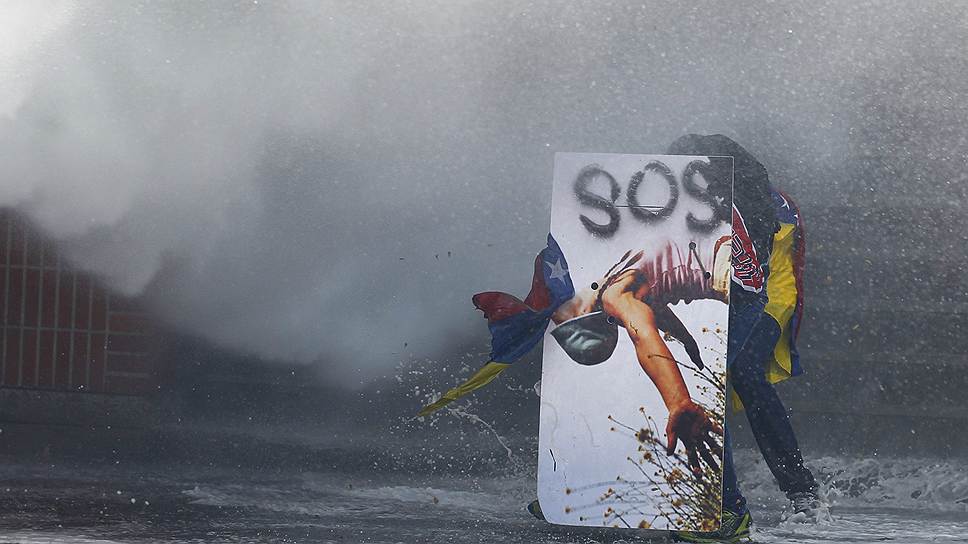 Демонстрант прячется от струи водомета во время беспорядков в Каракасе