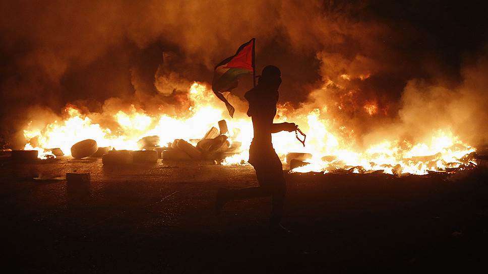 Палестинский протестующий размахивает палестинским флагом, проходя мимо горящих шин, которые остались после столкновений с израильскими войсками, — в знак протеста против израильского наступления в секторе Газа