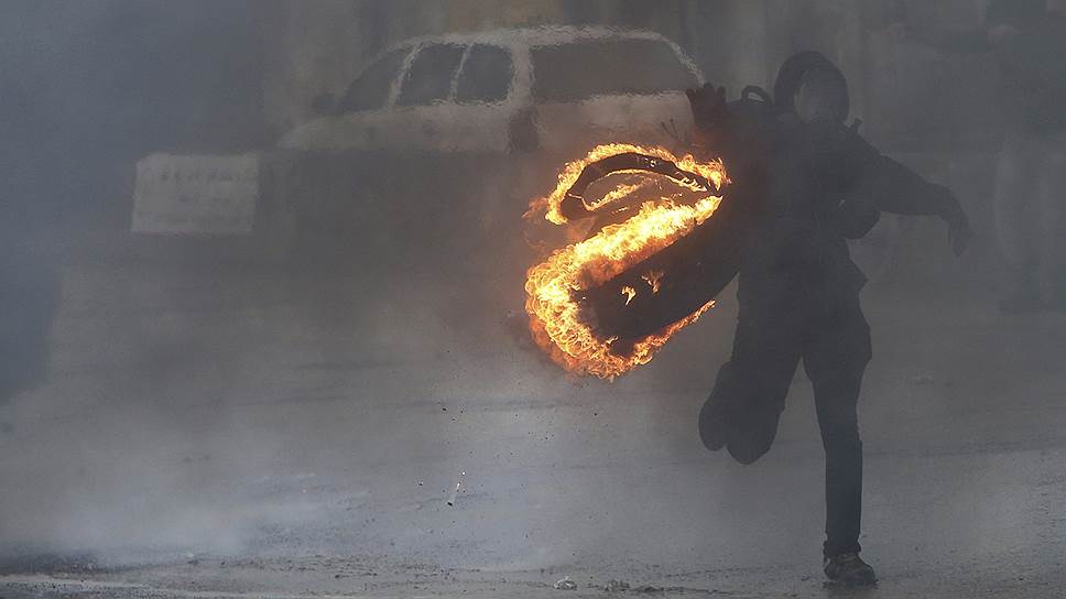 Протестующий палестинец в маске несет горящую шину во время столкновения с израильскими военными