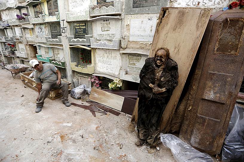 Гватемала, республика Гватемала. Чистильщик могил отдыхает возле мумифицированного тела, эксгумированного из-за истечения срока аренды захоронения