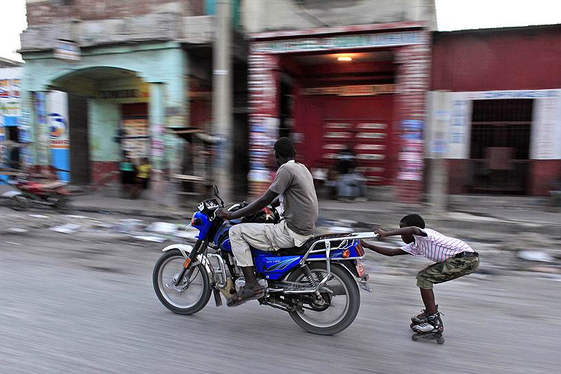 Порт-о-Пренс, Гаити. Ребенок едет на роликовых коньках, зацепившись за мотоцикл