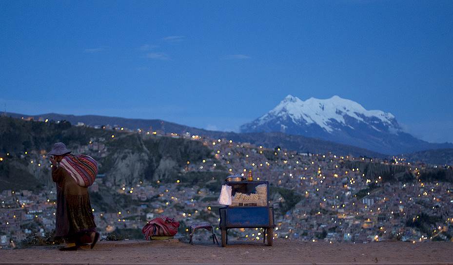 Лас-Пас, Боливия. Женщина идет мимо лотка с попкорном на фоне покрытой снегом горы Ильимани