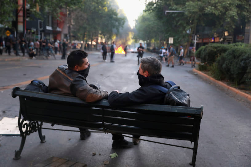 Сантьяго, Чили. Двое демонстрантов сидят на скамейке, пока происходят столкновения протестующих против правительства Чили с полицией