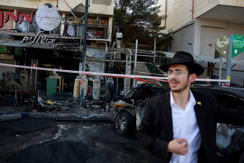 Мужчина на фоне здания, пострадавшего от ракетного удара, в израильском приграничном городе Кирьят-Шмона