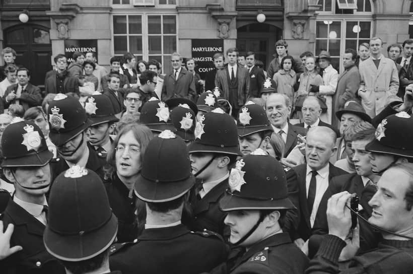 Джон Леннон и Йоко Оно (справа от него, ее не видно, заслоняет полицейский шлем) покидают здание Королевского суда лондонского района Марилебон — через день после обыска, проведенного в их квартире Норманом Пилчером