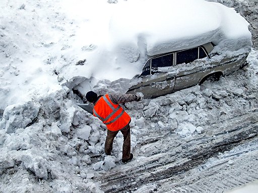 На снимке: дворник разгребает снег у припаркованного заснеженного автомобиля.