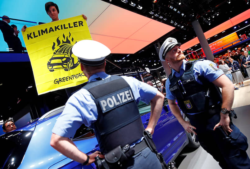 Офицеры полиции приглядывают за активистом «Гринпис», который разворачивает плакат про то, что автомобили — «убийцы климата»