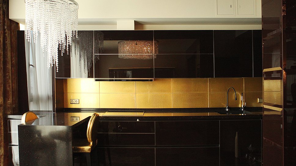Благодаря отражающим свойствам материала, из которого сделаны кухонные фасады, и золотой плитке, два функционально разных помещения — кухня и гостиная — соединяются в одно гармоничное целое